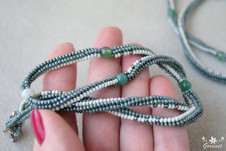 May include: A multi-strand necklace made with small, gray, white, and green beads. The necklace is made with a variety of beads, including round, oval, and square beads. The necklace is strung on a silver chain.
