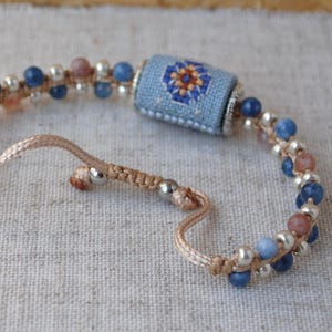 May include: Handmade bracelet featuring a central light blue cylinder with a floral cross-stitch design in orange, blue, and white. The bracelet includes blue and pink beads, silver-colored beads, and a beige cord. The bracelet is adjustable.
