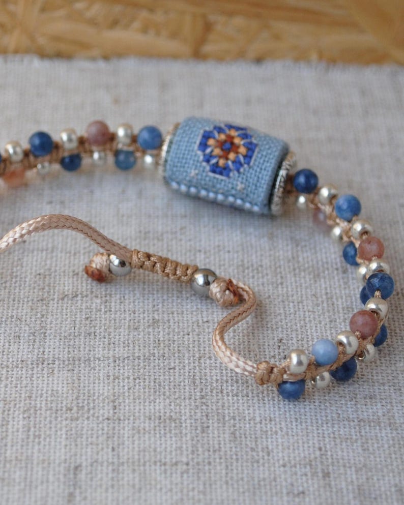 May include: Handmade bracelet featuring a central light blue cylinder with a floral cross-stitch design in orange, blue, and white. The bracelet includes blue and pink beads, silver-colored beads, and a beige cord. The bracelet is adjustable.