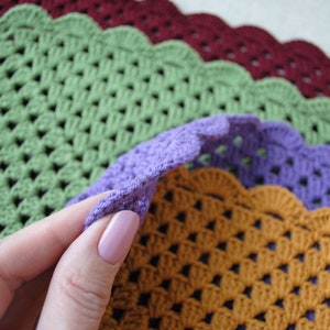 May include: Four crocheted placemats in different colors: green, purple, orange, and burgundy. The placemats have a scalloped edge and a woven pattern.
