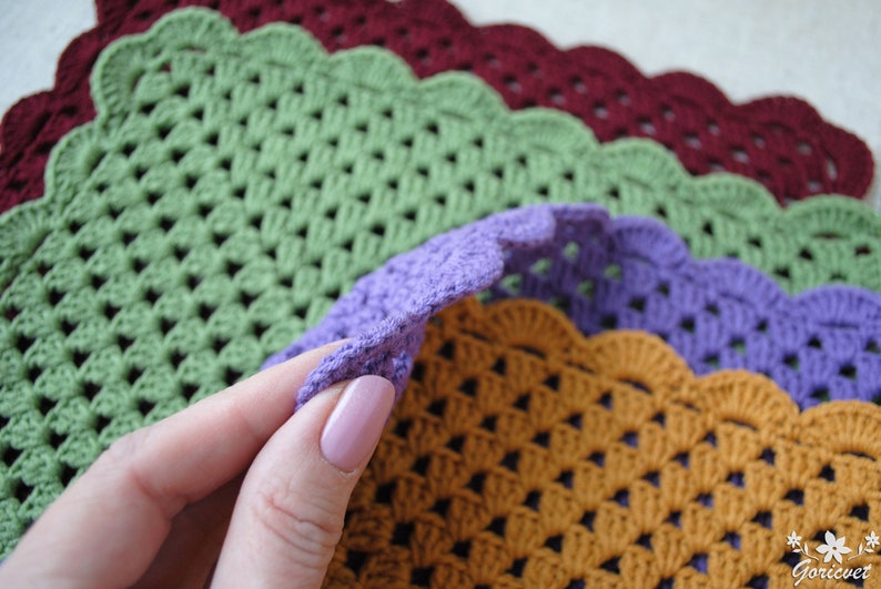 May include: Four crocheted placemats in different colors: green, purple, orange, and burgundy. The placemats have a scalloped edge and a woven pattern.