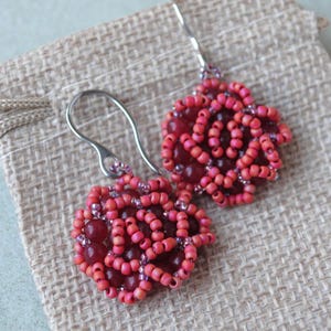 Peut inclure: Une paire de boucles d'oreilles faites à la main avec un motif floral. Les boucles d'oreilles sont composées de petites perles rondes rouges et roses, avec un centre rouge plus foncé. Les crochets d'oreilles sont argentés. Les boucles d'oreilles reposent sur un tissu beige.