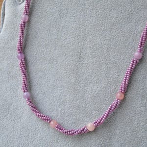 Puede incluir: Un collar delicado hecho con peque&ntilde;as cuentas moradas y cuentas rosadas m&aacute;s grandes. El collar se muestra sobre una superficie de terciopelo gris.