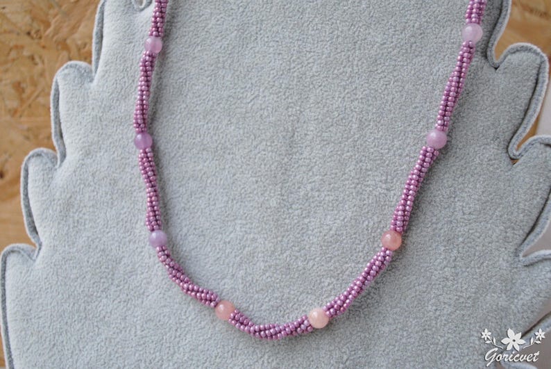 Puede incluir: Un collar delicado hecho con peque&ntilde;as cuentas moradas y cuentas rosadas m&aacute;s grandes. El collar se muestra sobre una superficie de terciopelo gris.