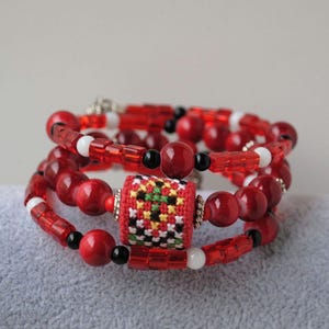 May include: A multi-strand wrap bracelet featuring red, white, and black beads. The design includes translucent red cylindrical beads, opaque red spherical beads, and small white and black accent beads. A central cylindrical bead features a cross-stitch pattern in red, yellow, green, and black thread.