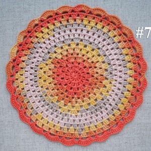 Puede incluir: Un tapete de ganchillo circular hecho a mano en una vibrante paleta de colores. El tapete presenta anillos conc&eacute;ntricos de color, que van desde un centro de coral c&aacute;lido hasta tonos de amarillo, gris y melocot&oacute;n, con un borde naranja festoneado. El n&uacute;mero "#7" es visible.