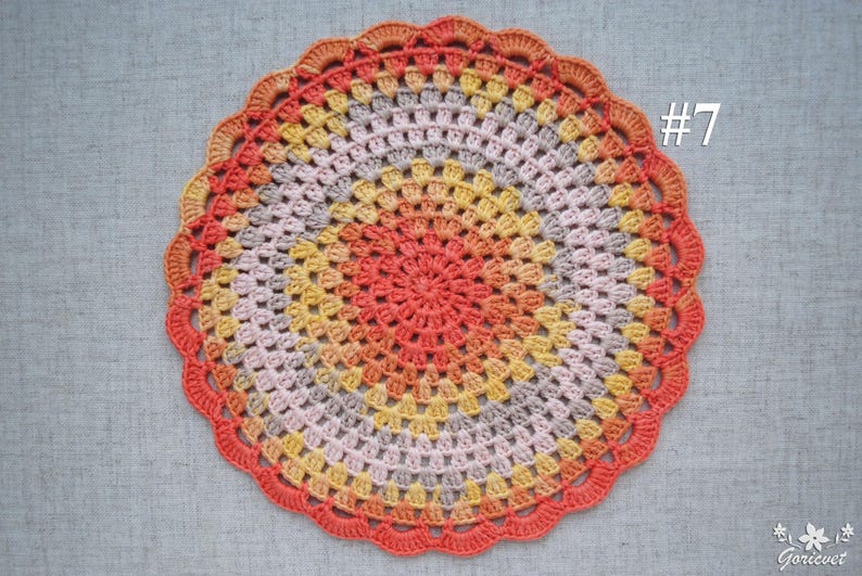 Puede incluir: Un tapete de ganchillo circular hecho a mano en una vibrante paleta de colores. El tapete presenta anillos conc&eacute;ntricos de color, que van desde un centro de coral c&aacute;lido hasta tonos de amarillo, gris y melocot&oacute;n, con un borde naranja festoneado. El n&uacute;mero "#7" es visible.