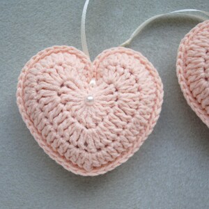 May include: Two pink crocheted hearts with white pearl accents hanging from white ribbon.
