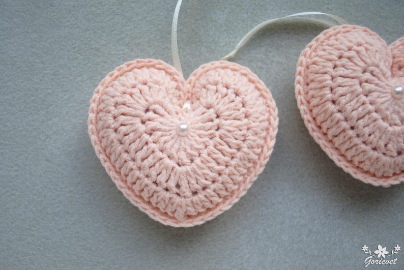 May include: Two pink crocheted hearts with white pearl accents hanging from white ribbon.