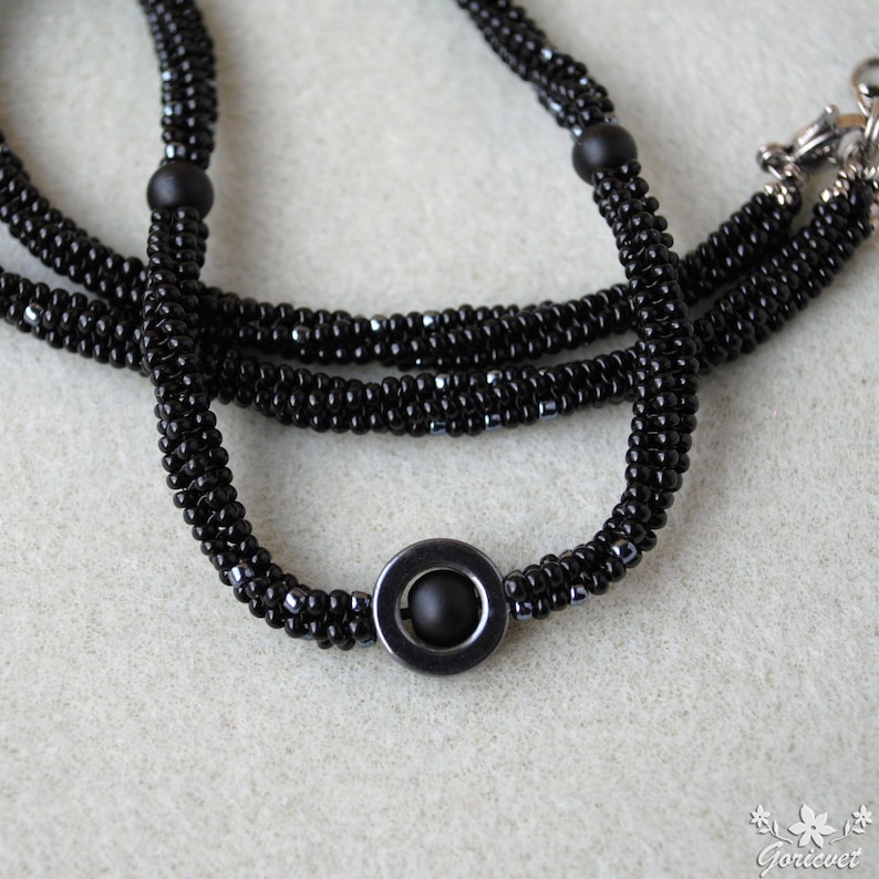 May include: A black beaded necklace with a round, black, metal pendant. The necklace is made of small, black beads and has a simple, elegant design.