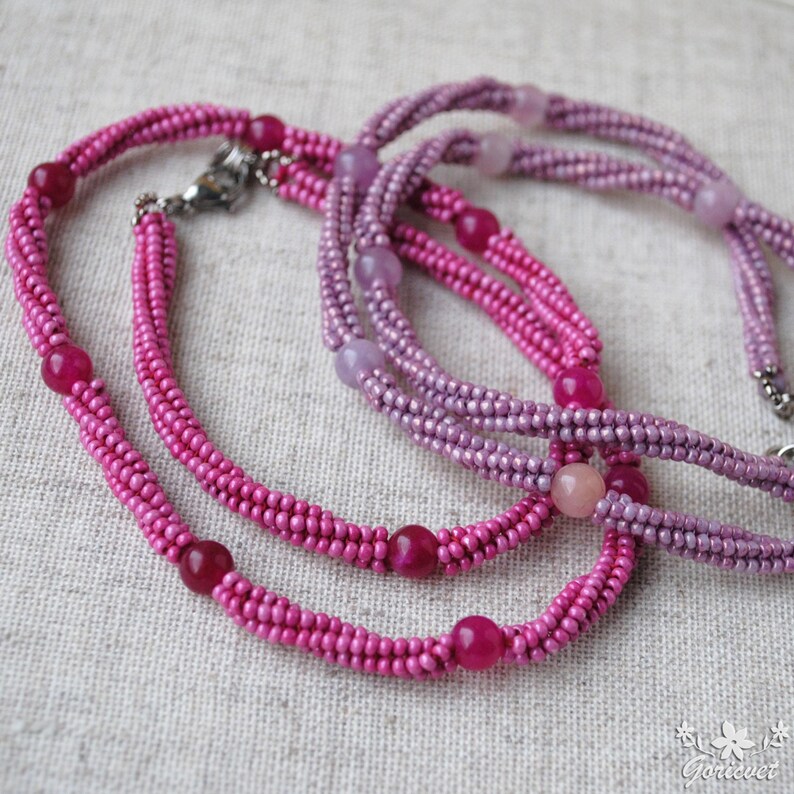 Puede incluir: Dos hileras de collares de cuentas rosadas y moradas con acentos de piedras preciosas redondas.