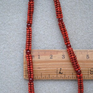 May include: A close-up of a red beaded necklace with a wooden ruler for scale. The necklace is made of small, round beads and interspersed with larger, darker red beads. The ruler shows measurements in inches.