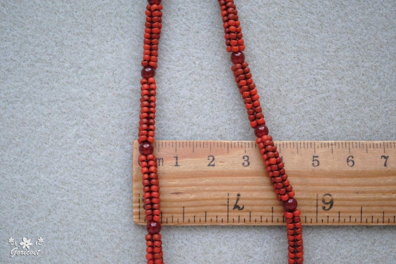 May include: A close-up of a red beaded necklace with a wooden ruler for scale. The necklace is made of small, round beads and interspersed with larger, darker red beads. The ruler shows measurements in inches.