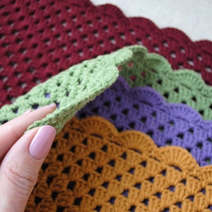 May include: Four crocheted placemats in different colors: burgundy, green, purple, and orange. The placemats have a scalloped edge and a woven pattern.