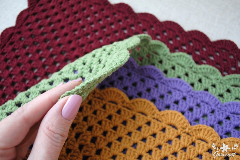 May include: Four crocheted placemats in different colors: burgundy, green, purple, and orange. The placemats have a scalloped edge and a woven pattern.