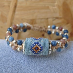 May include: Handmade bracelet featuring a central light blue embroidered cylinder with a floral design in blue, orange, and brown. The bracelet has blue and tan beads, with silver-colored accents, strung on a natural-colored cord.