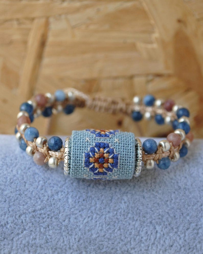 May include: Handmade bracelet featuring a central light blue embroidered cylinder with a floral design in blue, orange, and brown. The bracelet has blue and tan beads, with silver-colored accents, strung on a natural-colored cord.