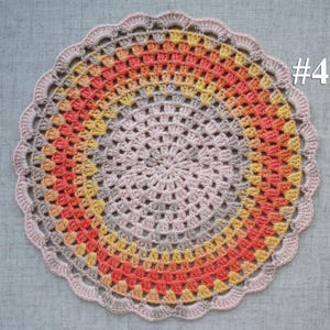 Puede incluir: Un tapete de ganchillo circular hecho a mano con un borde festoneado. El tapete presenta anillos conc&eacute;ntricos de color, que transicionan de un centro rosa claro a tonos de amarillo, naranja y gris. El n&uacute;mero "#4" es visible en la esquina superior derecha.