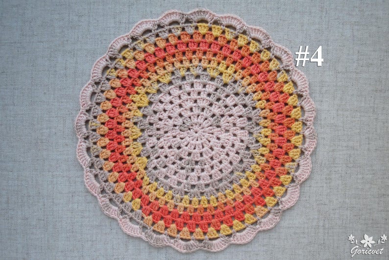 Puede incluir: Un tapete de ganchillo circular hecho a mano con un borde festoneado. El tapete presenta anillos conc&eacute;ntricos de color, que transicionan de un centro rosa claro a tonos de amarillo, naranja y gris. El n&uacute;mero "#4" es visible en la esquina superior derecha.
