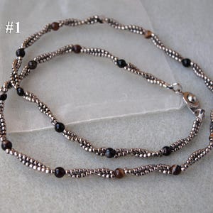 Op de afbeelding: Een zilveren ketting met zwarte en bruine kralen. De ketting is dubbelstrengs en heeft een sluiting. De tekst "Style #1" is zichtbaar in de afbeelding.