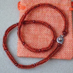 May include: A handcrafted necklace made of small, reddish-orange beads. The necklace is displayed on a textured orange pouch. The necklace has a silver clasp. The beads are a consistent size and shape, creating a smooth, elegant look.