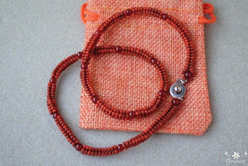 May include: A handcrafted necklace made of small, reddish-orange beads. The necklace is displayed on a textured orange pouch. The necklace has a silver clasp. The beads are a consistent size and shape, creating a smooth, elegant look.