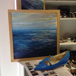 May include: A framed painting of a blue ocean with a light blue sky and clouds. A pair of blue patent leather shoes with gold buckles are in the foreground.