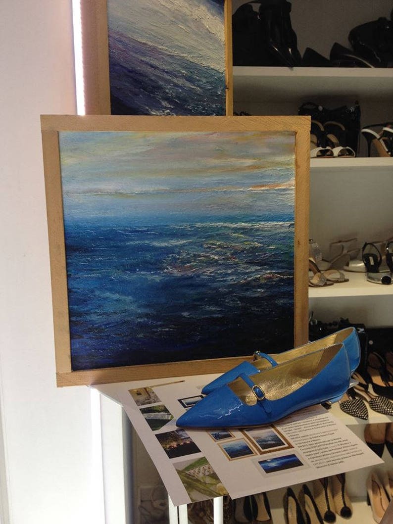 May include: A framed painting of a blue ocean with a light blue sky and clouds. A pair of blue patent leather shoes with gold buckles are in the foreground.