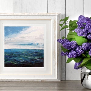 May include: A white framed painting of a seascape with blue and green water and a cloudy sky. A bouquet of purple lilac flowers in a silver vase is on a wooden table next to the painting.