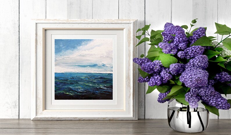 May include: A white framed painting of a seascape with blue and green water and a cloudy sky. A bouquet of purple lilac flowers in a silver vase is on a wooden table next to the painting.
