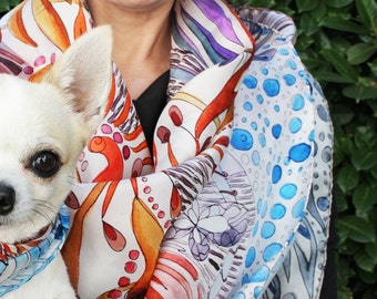 May include: A white chihuahua dog wearing a blue and white patterned bandana is being held by a person wearing a colorful silk scarf with a white background. The scarf has a variety of patterns, including orange, blue, and purple.
