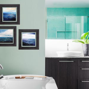 May include: Three framed seascape paintings in a bathroom setting. The paintings are in black frames and feature blue and white water with clouds in the sky. The bathroom has a white sink and a white bathtub.
