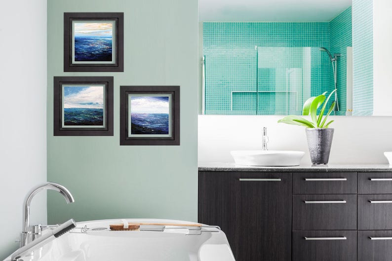 May include: Three framed seascape paintings in a bathroom setting. The paintings are in black frames and feature blue and white water with clouds in the sky. The bathroom has a white sink and a white bathtub.