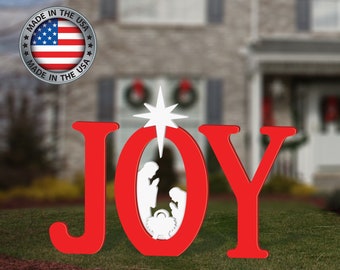 Peut inclure: Décoration de Noël extérieure rouge avec le mot "JOY" et une étoile blanche et une scène de la nativité. La décoration est sur une pelouse verte avec une maison en arrière-plan. Un emblème "Made in the USA" est en haut à gauche.