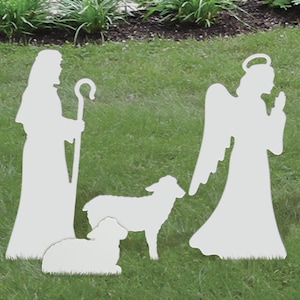 May include: White silhouette cutout of a shepherd, an angel, and two sheep. The figures are standing on a green grassy lawn.