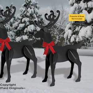 May include: Two black metal reindeer yard decorations with red bows. The reindeer are standing in a snowy yard with evergreen trees in the background. The text "Presents & Bows Not Included" is on a yellow sign.