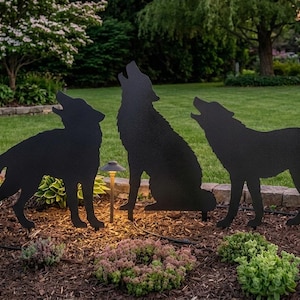 May include: A set of three black metal silhouettes of wolves in a howling pose, positioned upright in a garden bed. The figures depict two wolves standing and one wolf sitting, all facing upward with their heads tilted back in a classic howling stance.