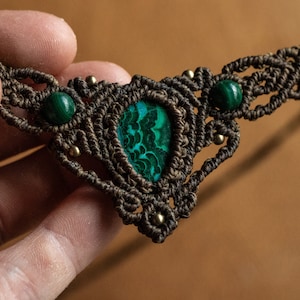 May include: A brown macrame necklace with a green malachite stone pendant and two smaller green malachite stones on the sides.