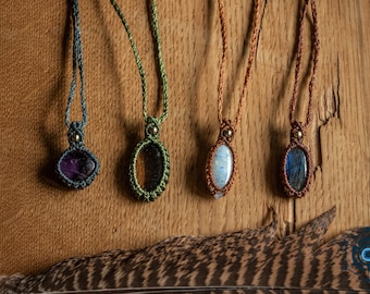 May include: Four macrame necklaces with oval-shaped gemstone pendants. The gemstones are purple, green, white, and blue. Each necklace has a different colored cord, and a small gold-colored bead at the top of the pendant.