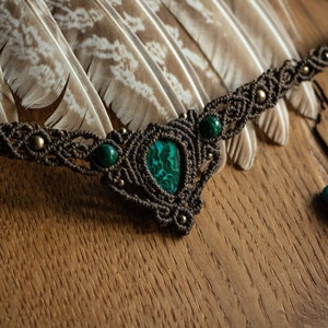 May include: A brown macrame necklace with a green malachite stone pendant and smaller green stones. The necklace is displayed on a wooden surface with feathers and malachite stones.
