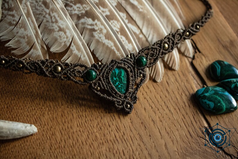 May include: A brown macrame necklace with a green malachite stone pendant and smaller green stones. The necklace is displayed on a wooden surface with feathers and malachite stones.