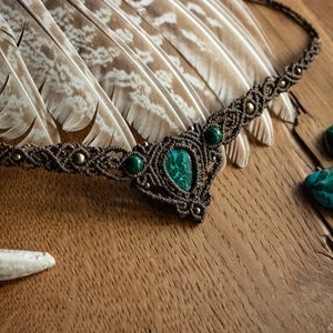 May include: A brown macrame necklace with a green malachite stone pendant and smaller green stones. The necklace is on a wooden surface with feathers and a piece of antler.