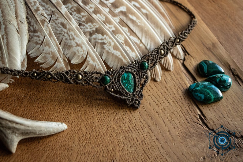 May include: A brown macrame necklace with a green malachite stone pendant and smaller green stones. The necklace is on a wooden surface with feathers and a piece of antler.