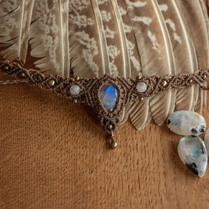 May include: A brown macrame choker necklace with a large iridescent moonstone pendant and smaller moonstone accents. The necklace is displayed on a wooden surface with feathers and loose moonstones.