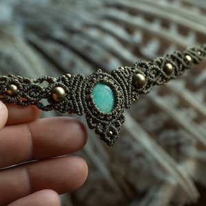 Peut inclure: Un collier avec une pierre précieuse verte, des perles dorées et un motif en macramé.