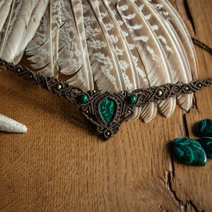 May include: A brown macrame necklace with a green malachite stone pendant and smaller green stones on the sides. The necklace is displayed on a wooden surface with feathers and antlers.
