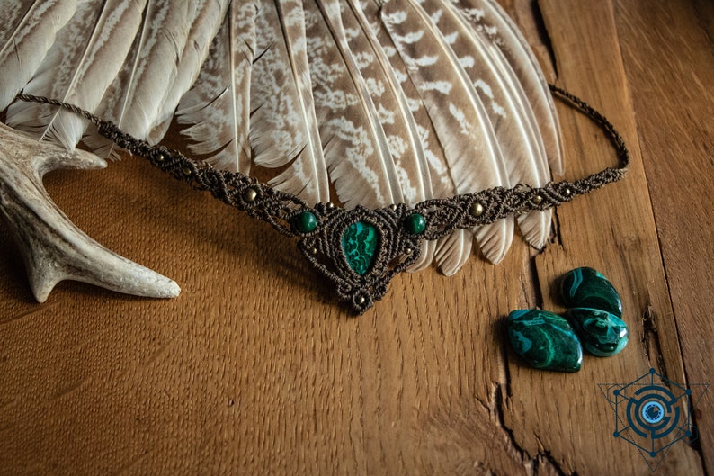 May include: A brown macrame necklace with a green malachite stone pendant and smaller green stones on the sides. The necklace is displayed on a wooden surface with feathers and antlers.