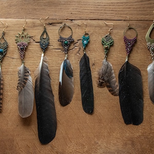 Op de afbeelding: Negen paar veren oorbellen met macramé en edelsteen accenten. De oorbellen zijn in verschillende kleuren, waaronder groen, blauw, paars en bruin. De veren zijn zwart, bruin en wit.