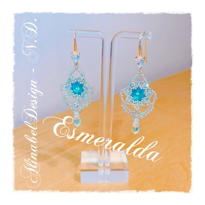Könnte beinhalten: Ein Paar silberne Ohrringe mit einem türkisblauen Edelstein in der Mitte jedes Ohrrings. Die Ohrringe hängen an einem durchsichtigen Acrylständer. Der Text "AlinaBel Design - N.D. Esmeralda" ist in Kursivschrift auf dem Bild geschrieben.