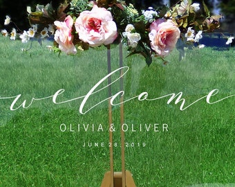 May include: Clear acrylic wedding welcome sign with white calligraphy lettering that reads "welcome Olivia & Oliver June 28, 2019". The sign is decorated with pink and white flowers.